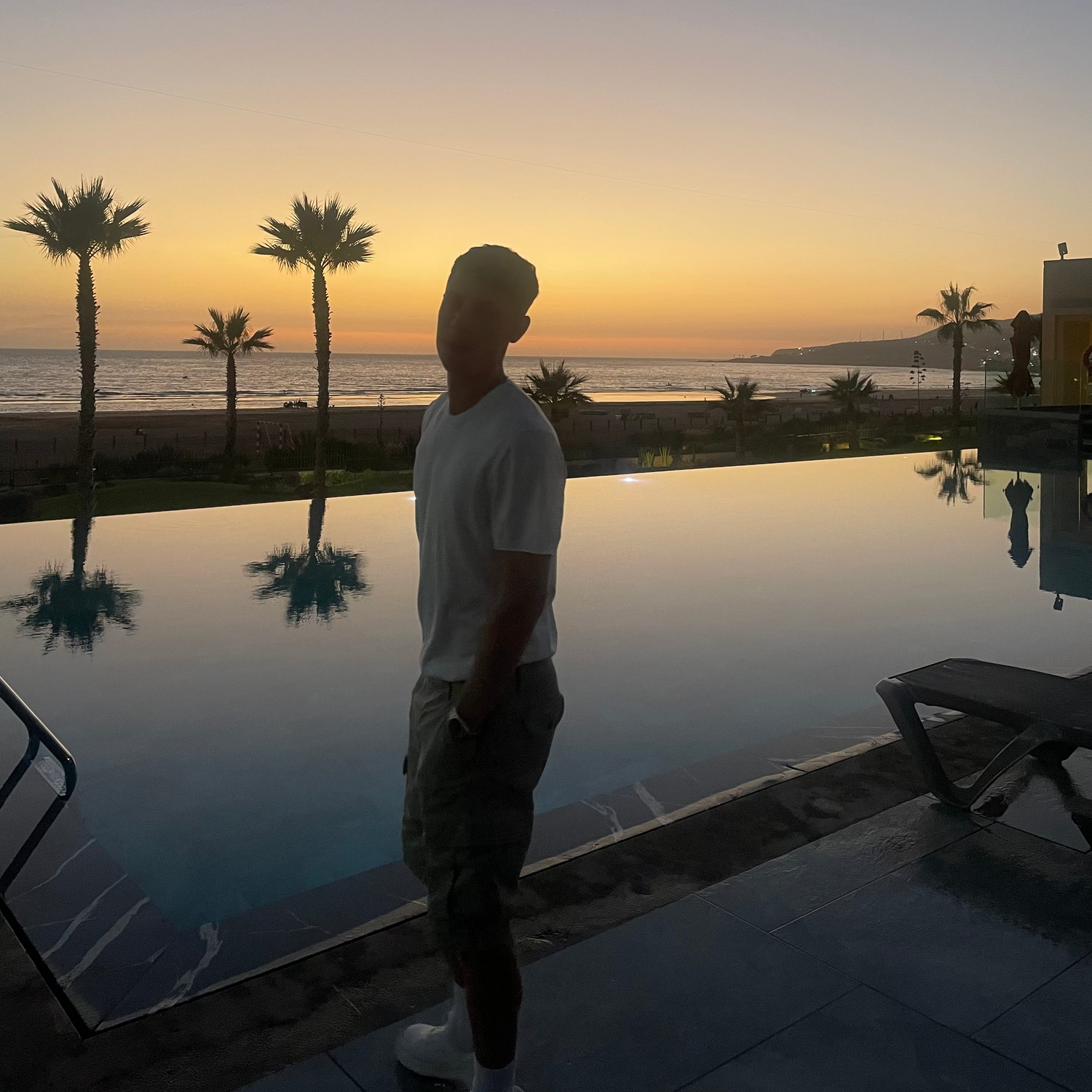 Person standing by a pool with palm trees and sunset in the background