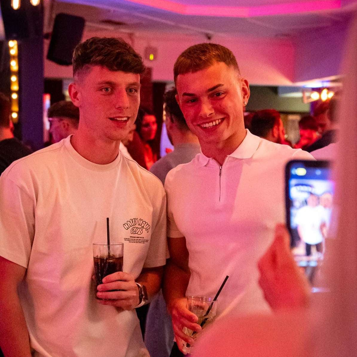 Two young men posing together at a party with drinks, surrounded by pink and purple lighting.